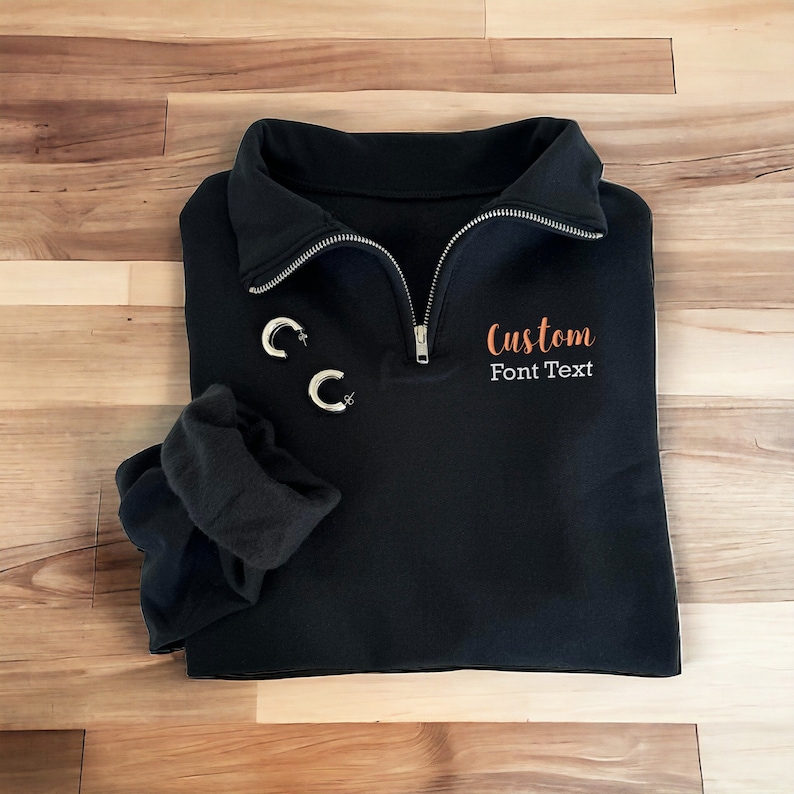 May include: Black quarter-zip sweatshirt with a custom orange embroidered font text that says "Custom Font Text".