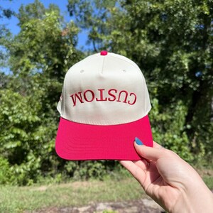 May include: A cream-colored baseball cap with a bright pink brim. The word "CUSTOM" is embroidered in red across the front. The cap has a red button on top.