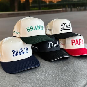May include: A collection of baseball caps with various embroidered titles, including "DAD", "GRANDPA", and "PAPA". The caps feature contrasting brims in navy, black, green, and red, with the text in different fonts and colors.