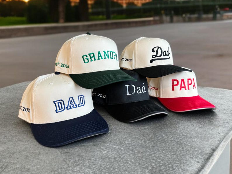 May include: A collection of baseball caps with various embroidered titles, including "DAD", "GRANDPA", and "PAPA". The caps feature contrasting brims in navy, black, green, and red, with the text in different fonts and colors.
