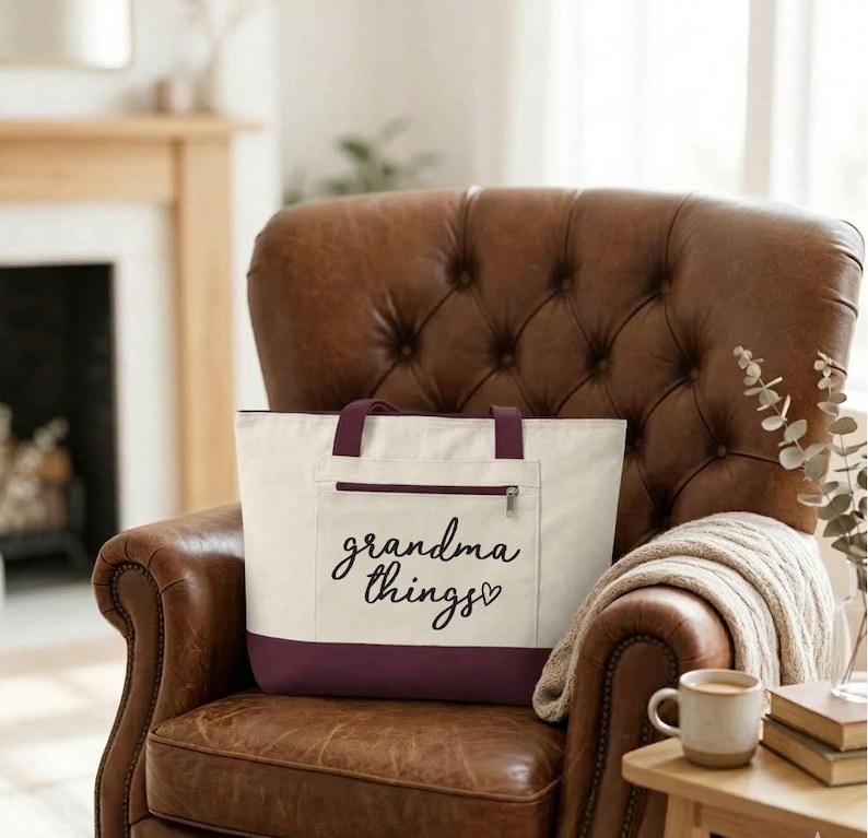 Op de afbeelding: Cr&egrave;mekleurige draagtas met een bordeauxrode basis en handvatten, met een voorvak met ritssluiting. De woorden "grandma things" staan in cursief op de voorkant. De tas staat op een bruine leren fauteuil.