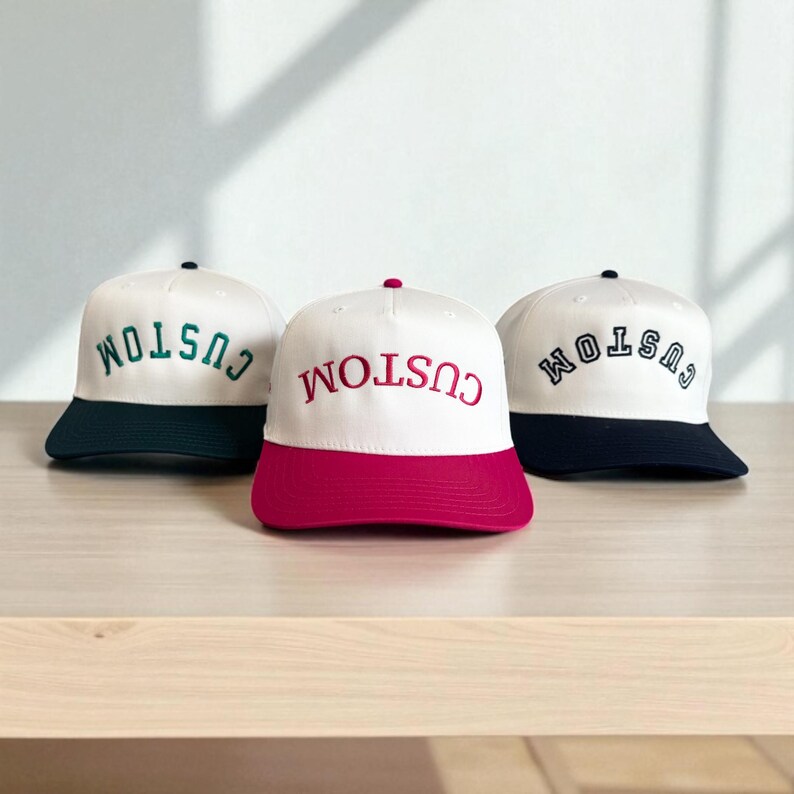 May include: Three white baseball caps with the word "CUSTOM" embroidered in different colors. The caps have colored brims in dark green, red, and navy blue. The caps are displayed on a light brown surface.