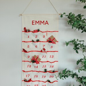 May include: A cream-colored advent calendar with red accents and the name "EMMA" printed at the top. The calendar has 24 pockets, each numbered and decorated with small gifts and greenery. It hangs from a wooden dowel.