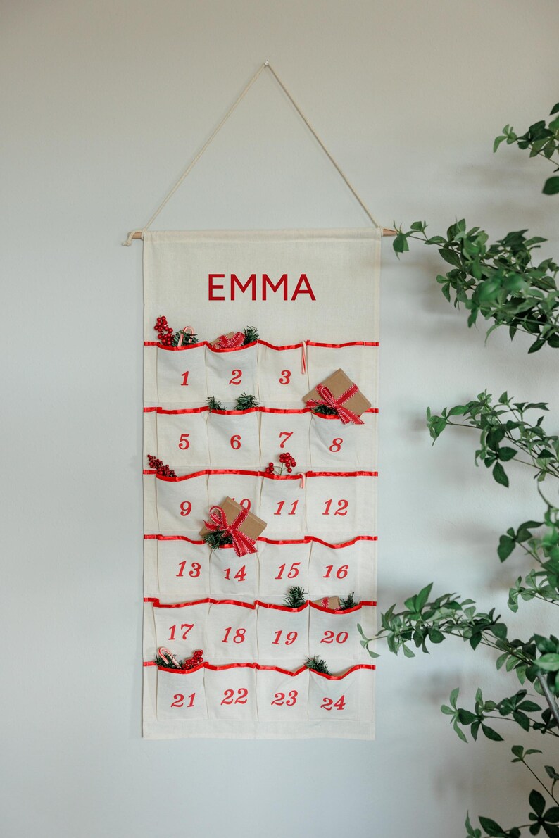 May include: A cream-colored advent calendar with red accents and the name "EMMA" printed at the top. The calendar has 24 pockets, each numbered and decorated with small gifts and greenery. It hangs from a wooden dowel.