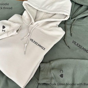 May include: Sand-colored hoodie with black thread, featuring a hood and drawstring. The sleeve displays a heart and the letter "Z". The front of the hoodie has the roman numerals "VII.XXII.MMXX". A heather dark green hoodie is also shown.