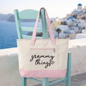 Op de afbeelding: Cr&egrave;mekleurige draagtas met roze accenten, waaronder de bodem, handvatten en een voorvak. De tekst "grammy things" is in zwart schrift gedrukt. De tas staat op een lichtblauwe stoel.