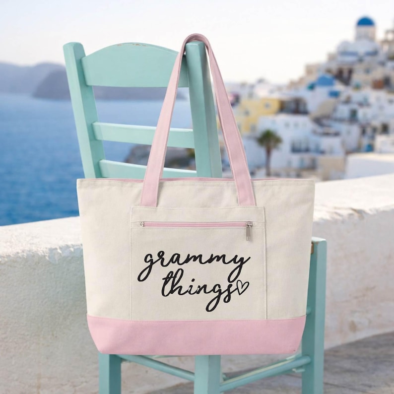 Op de afbeelding: Cr&egrave;mekleurige draagtas met roze accenten, waaronder de bodem, handvatten en een voorvak. De tekst "grammy things" is in zwart schrift gedrukt. De tas staat op een lichtblauwe stoel.