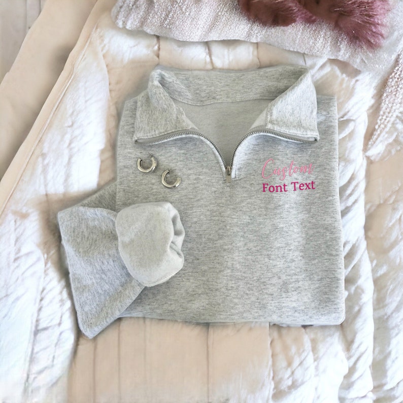 May include: A grey sweatshirt with a pink embroidered design that says "Capricorn" with the text "Font Text" underneath. The sweatshirt has a zip and a mock neck.
