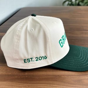 May include: Cream-coloured baseball cap with a dark green brim. The word "GRA" is embroidered in green on the front, with "EST. 2016" on the side. The cap has a dark green button on top.