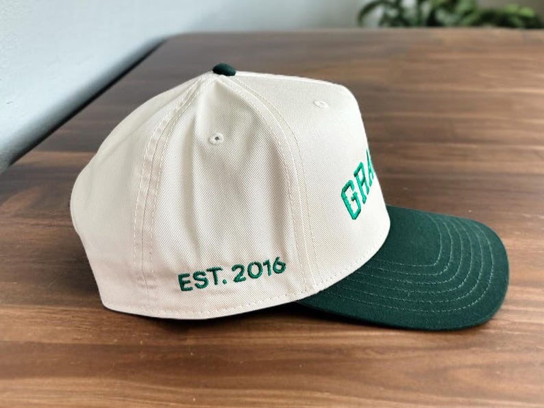 May include: Cream-coloured baseball cap with a dark green brim. The word "GRA" is embroidered in green on the front, with "EST. 2016" on the side. The cap has a dark green button on top.