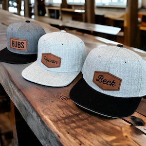 May include: Three gray baseball caps with black brims are displayed on a wooden surface. Each cap features a brown leather patch with a name: "BUBS", "Rowan", and "Beck". The caps have a classic, casual style.