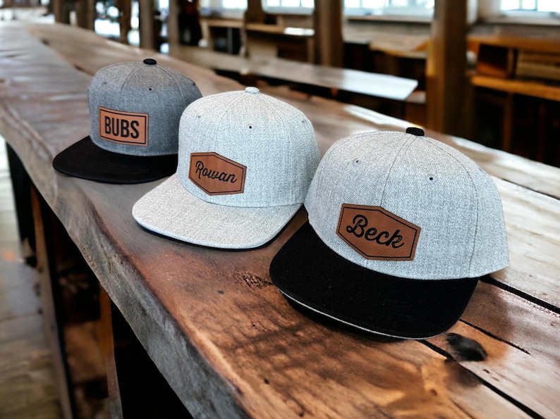 May include: Three gray baseball caps with black brims are displayed on a wooden surface. Each cap features a brown leather patch with a name: "BUBS", "Rowan", and "Beck". The caps have a classic, casual style.