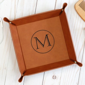 May include: A brown leather catchall tray with a stitched edge and a monogrammed initial "M" in a circle.
