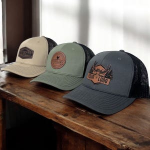 May include: Three trucker hats with mesh backs and curved visors are displayed on a wooden surface. Each hat features a different logo patch: a mountain scene, a rooster, and a deer with trees. The text "YOUR LOGO" is included on each patch.