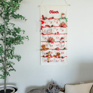 May include: A cream-colored Christmas advent calendar with red accents and the name "Oliver" printed at the top. The calendar has 24 pockets, each with a number and a small holiday-themed toy or decoration. A dog stands near the calendar.