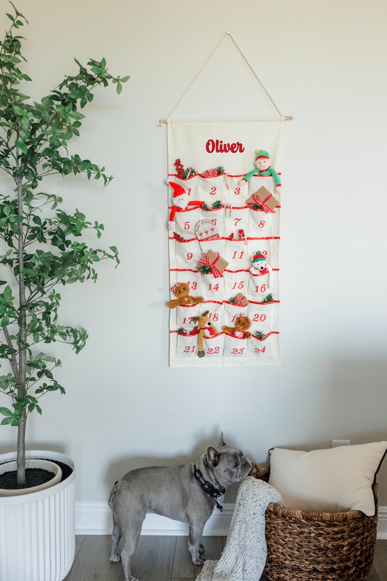 May include: A cream-colored Christmas advent calendar with red accents and the name "Oliver" printed at the top. The calendar has 24 pockets, each with a number and a small holiday-themed toy or decoration. A dog stands near the calendar.