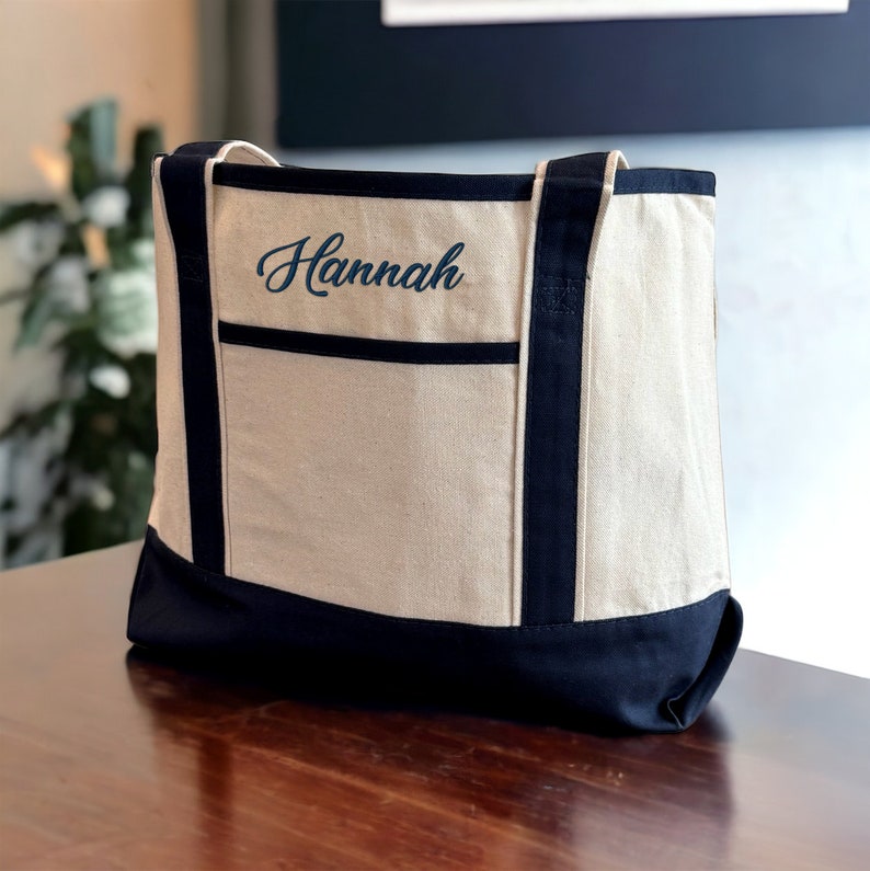 May include: A beige canvas tote bag with black trim and a black bottom. The bag has a pocket on the front and the name "Hannah" is embroidered in blue on the front.