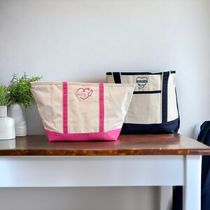 May include: Two large canvas tote bags with pink and black handles. The bag on the left has the name "Bailey" embroidered on it with a stethoscope inside a heart. The bag on the right has the name "Natalie" embroidered on it with a stethoscope inside a heart.