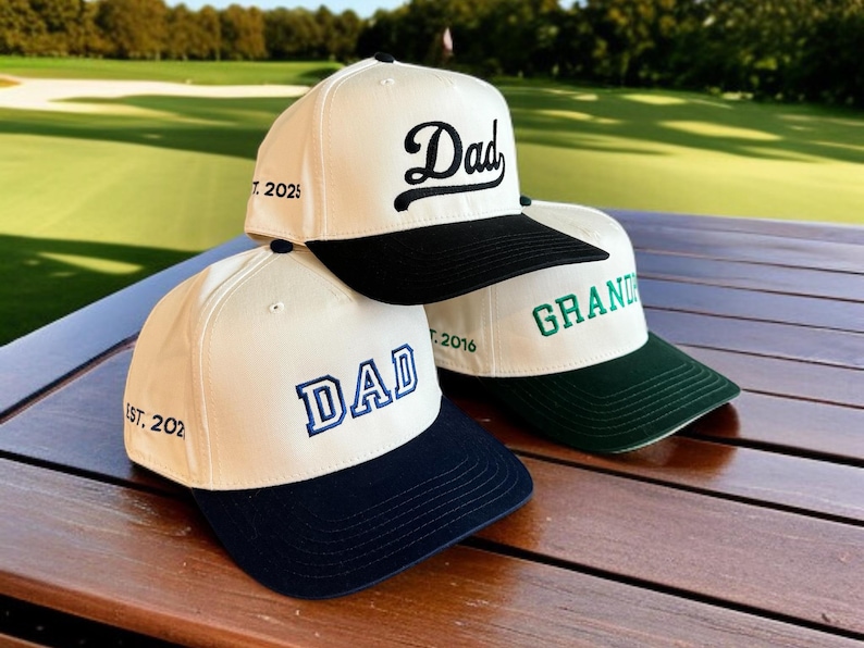 May include: Three baseball caps with white crowns and colored brims are stacked on a wooden surface. The caps feature the words "Dad" and "Grand" embroidered in various fonts and colors. The caps also have the years "2025" and "2016" embroidered on them.
