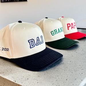 May include: Three baseball caps with white crowns and colored brims. The first cap has a navy blue brim and the word "DAD" embroidered in blue. The second cap has a green brim and the word "GRAND" embroidered in green. The third cap has a red brim and the word "PAP" embroidered in red.