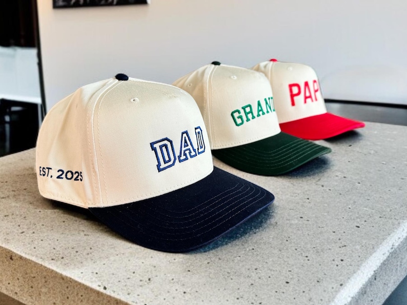 May include: Three baseball caps with white crowns and colored brims. The first cap has a navy blue brim and the word "DAD" embroidered in blue. The second cap has a green brim and the word "GRAND" embroidered in green. The third cap has a red brim and the word "PAP" embroidered in red.