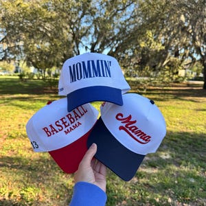 May include: Three baseball caps are displayed, each with a white crown and a coloured brim. One cap features the word "Mommin'" in blue, another says "Baseball Mama" with the number 13, and the third says "Mama" in red.