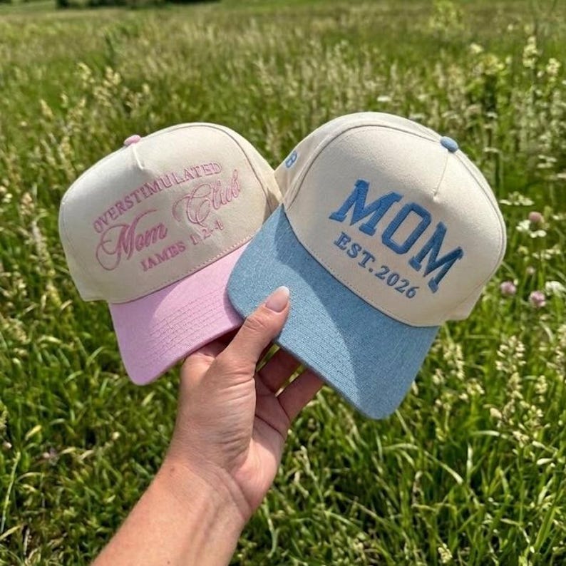 May include: Two baseball caps. One is beige with a pink peak and the words "OVERSTIMULATED Mom Club" in pink script. The other is beige with a blue peak and the word "MOM EST. 2026" in blue.