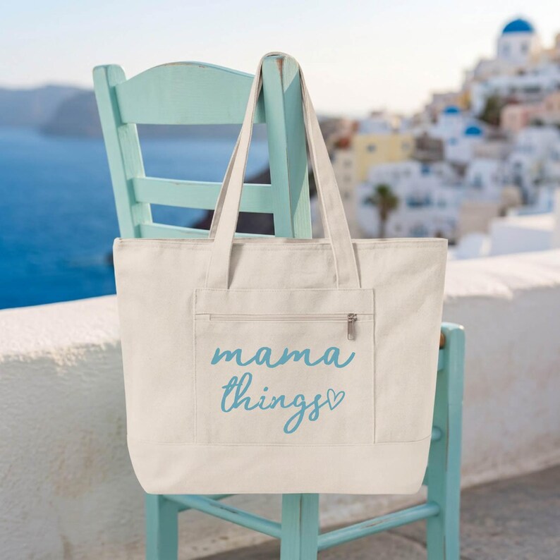 Op de afbeelding: Een cr&egrave;mekleurige draagtas met de woorden "mama things" in lichtblauw schrift. De tas heeft een voorvak met een rits en twee stevige handvatten. De tas hangt aan een lichtblauwe stoel.