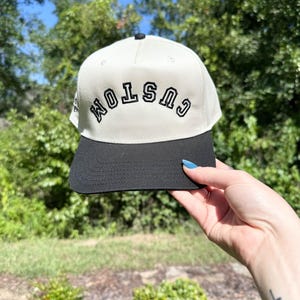 May include: A cream-colored baseball cap with a black brim and a black button on top. The word "CUSTOM" is embroidered in black on the front of the cap. The cap is held up against a background of green foliage and a blue sky.