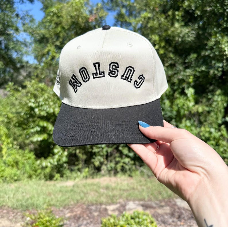 May include: A cream-colored baseball cap with a black brim and a black button on top. The word "CUSTOM" is embroidered in black on the front of the cap. The cap is held up against a background of green foliage and a blue sky.