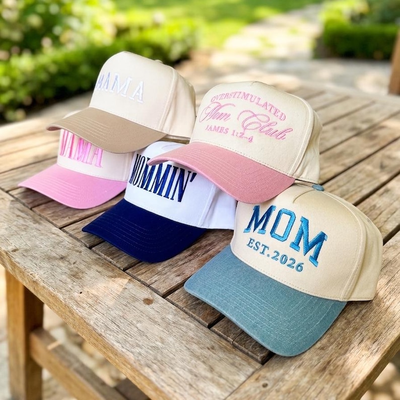 May include: Several baseball caps in shades of beige, pink, white, and blue. Each cap has embroidered text, including "MAMA," "MOM," and "OVERSTIMULATED Mom Club." The caps are arranged on a wooden table, showcasing a variety of designs.