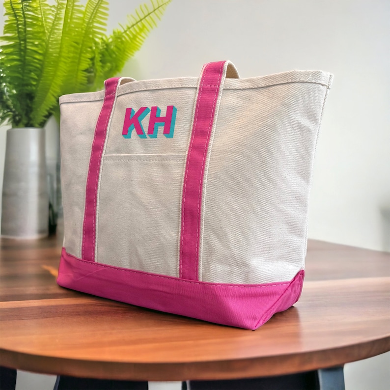 May include: A white canvas tote bag with pink trim and handles. The bag has a large pocket on the front and is monogrammed with the letters "KH" in turquoise and pink.