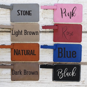May include: Eight leather luggage tags in various colours with text. The colours are stone, light brown, natural, dark brown, pink, rose, blue, and black. The text on the tags is the colour name.