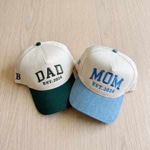 May include: Two baseball caps with white crowns and coloured brims. One cap has a green brim with the word "DAD" embroidered in dark green, and the text "EST.2026". The other cap has a light blue brim with the word "MOM" embroidered in blue, and the text "EST.2026".