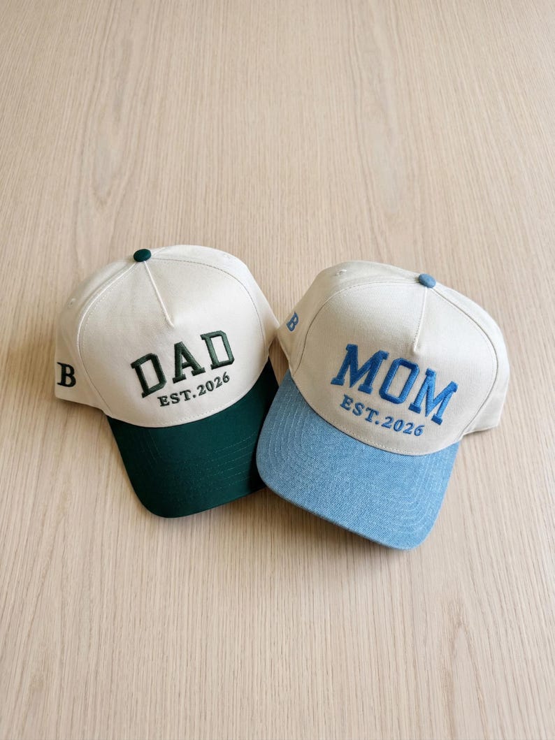 May include: Two baseball caps with white crowns and coloured brims. One cap has a green brim with the word "DAD" embroidered in dark green, and the text "EST.2026". The other cap has a light blue brim with the word "MOM" embroidered in blue, and the text "EST.2026".