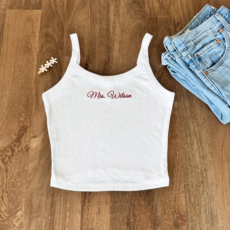 May include: White tank top with thin straps, featuring the embroidered text "Mrs. Wilson" in a burgundy script. The top is paired with blue jeans and a decorative hair clip on a wooden surface.