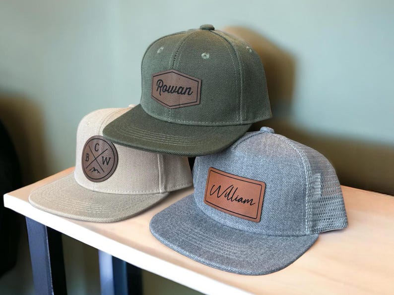 May include: Three personalized baseball caps with leather patches. The top cap is olive green with "Rowan" on a brown patch. The middle cap is tan with a circular patch. The bottom cap is gray with "William" on a brown patch.