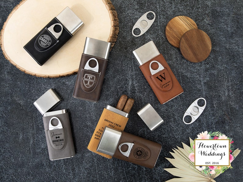 May include: A set of six personalized cigar cases with a built-in cutter. The cases are made of brown, black, and gray leather with silver metal accents. The cases are engraved with various names and messages, including "Groom Crew 2022", "Best Man", and "Groomsman".