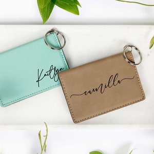 May include: Two personalized keychains. One is a light teal color with the name "Kaitlyn" in black script. The other is tan with the name "camilla" in black script. Both have a silver key ring.