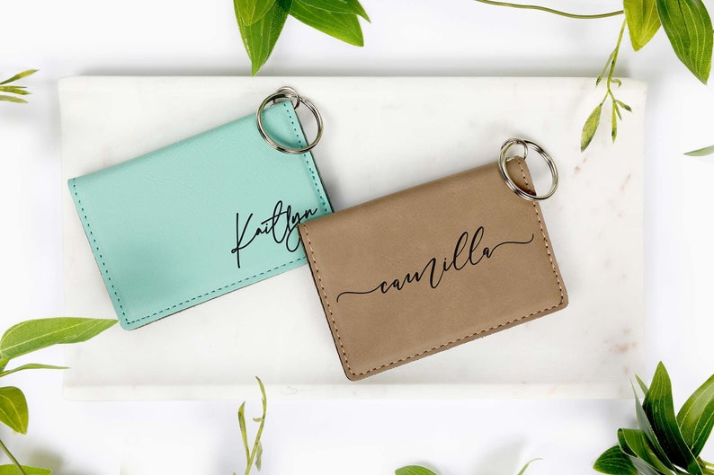 May include: Two personalized keychains. One is a light teal color with the name "Kaitlyn" in black script. The other is tan with the name "camilla" in black script. Both have a silver key ring.