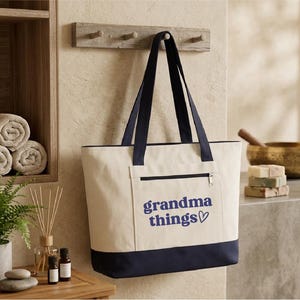 Op de afbeelding: Cr&egrave;mekleurige draagtas met marineblauwe accenten en handvatten. De tas heeft een voorvak met ritssluiting en de woorden "grandma things" in marineblauwe letters met een hartsymbool. Hij hangt aan een houten rek.