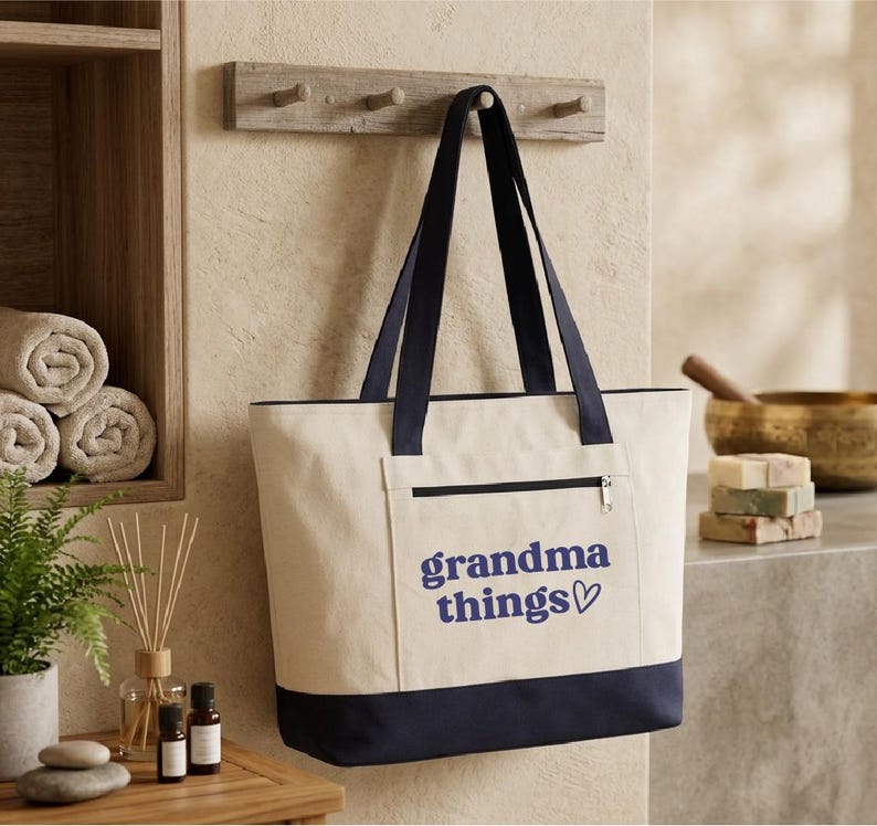 Op de afbeelding: Cr&egrave;mekleurige draagtas met marineblauwe accenten en handvatten. De tas heeft een voorvak met ritssluiting en de woorden "grandma things" in marineblauwe letters met een hartsymbool. Hij hangt aan een houten rek.