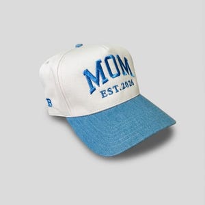 May include: A white and blue baseball cap featuring the embroidered word "MOM" in blue, with "EST.2026" underneath. The brim is light blue. The cap has a curved visor and a small "B" embroidered on the side.