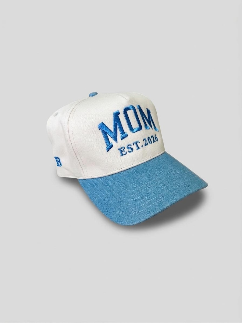 May include: A white and blue baseball cap featuring the embroidered word "MOM" in blue, with "EST.2026" underneath. The brim is light blue. The cap has a curved visor and a small "B" embroidered on the side.