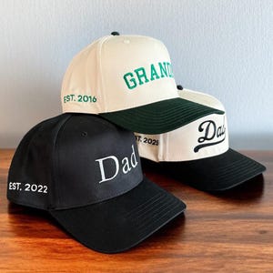 May include: Three baseball caps with embroidered text. One black cap says "Dad" with "EST. 2022". A cream-colored cap says "GRAND" with "EST. 2016". Another cream cap says "Dad" with "EST. 2025". All have curved brims.