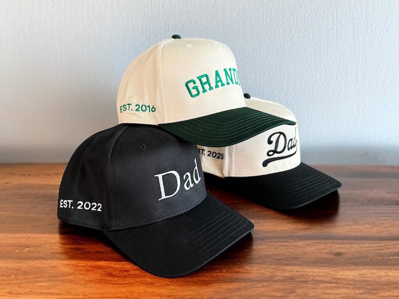 May include: Three baseball caps with embroidered text. One black cap says "Dad" with "EST. 2022". A cream-colored cap says "GRAND" with "EST. 2016". Another cream cap says "Dad" with "EST. 2025". All have curved brims.