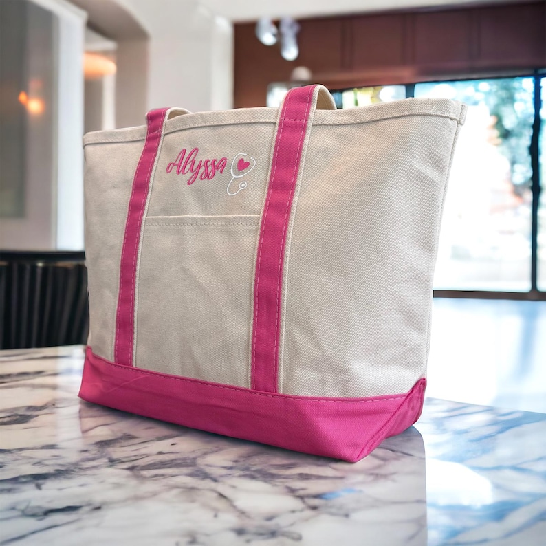 May include: A white canvas tote bag with pink trim and handles. The bag has a large pocket on the front and is embroidered with the name "Alyssa" and a pink heart with a stethoscope.