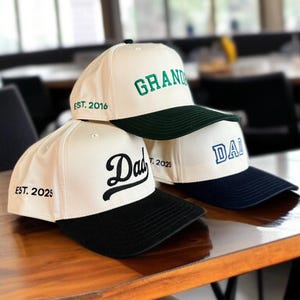 May include: Three baseball caps with white crowns and colored brims are stacked on a wooden surface. The top cap has "GRAND" in green, the middle cap has "Dad" in black, and the bottom cap has "DAD" in blue. Each cap has a different year embroidered on the side.