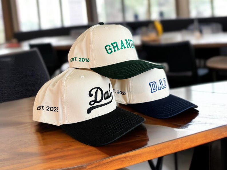 May include: Three baseball caps with white crowns and colored brims are stacked on a wooden surface. The top cap has "GRAND" in green, the middle cap has "Dad" in black, and the bottom cap has "DAD" in blue. Each cap has a different year embroidered on the side.