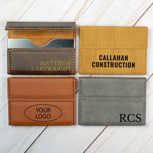 May include: Four different business card holders with various colors and materials. The first is brown leather with the name "Matthew Cartwright" in gold lettering. The second is a light brown wood with the text "CALLAHAN CONSTRUCTION" in black lettering. The third is a brown leather with the text "YOUR LOGO" in an oval shape. The fourth is a gray leather with the initials "RCS" in black lettering.